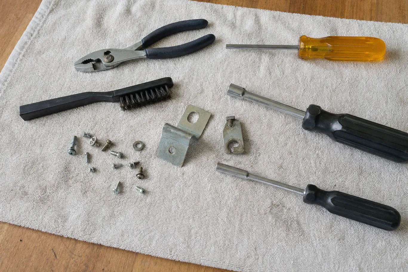 Slip-joint pliers, screwdrivers and a wire brush on a towel beside cam plates, washers and loose screws